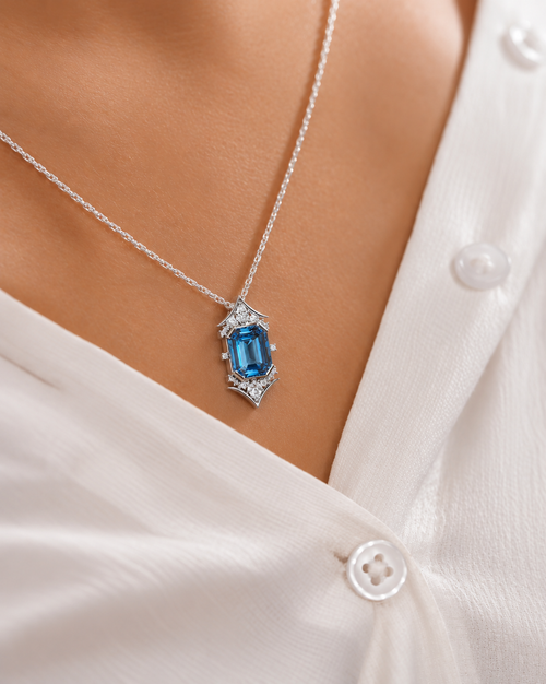 A close-up of a silver necklace with an emerald-cut blue gemstone pendant, worn on a model's neck over a white button-down shirt.