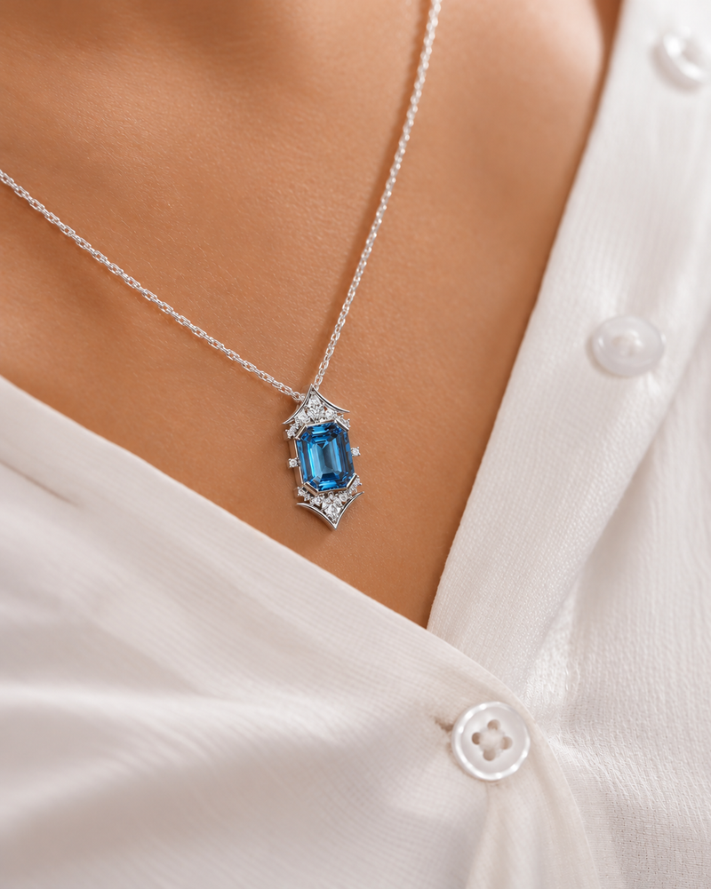 A close-up of a silver necklace with an emerald-cut blue gemstone pendant, worn on a model's neck over a white button-down shirt.