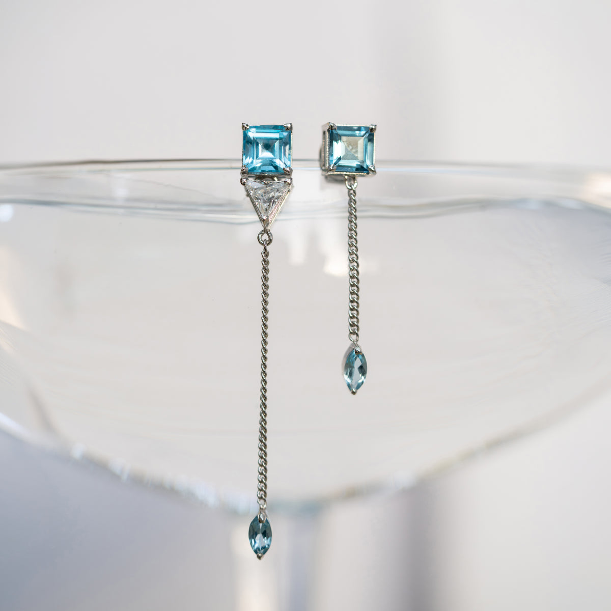 A pair of silver drop earrings with square light blue gemstones, a small triangular clear stone, and a dangling chain ending in a marquise-cut light blue gemstone, displayed on a clear glass surface against a white background.