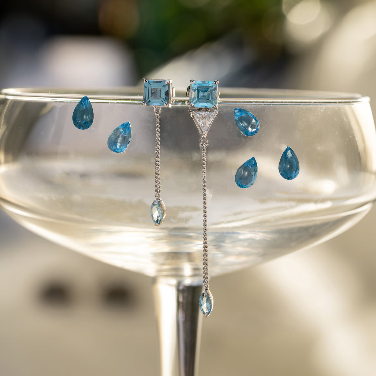 A pair of silver drop earrings with square blue gemstones and dangling teardrop-shaped blue stones, displayed draped over the rim of a clear coupe glass against a softly blurred background.